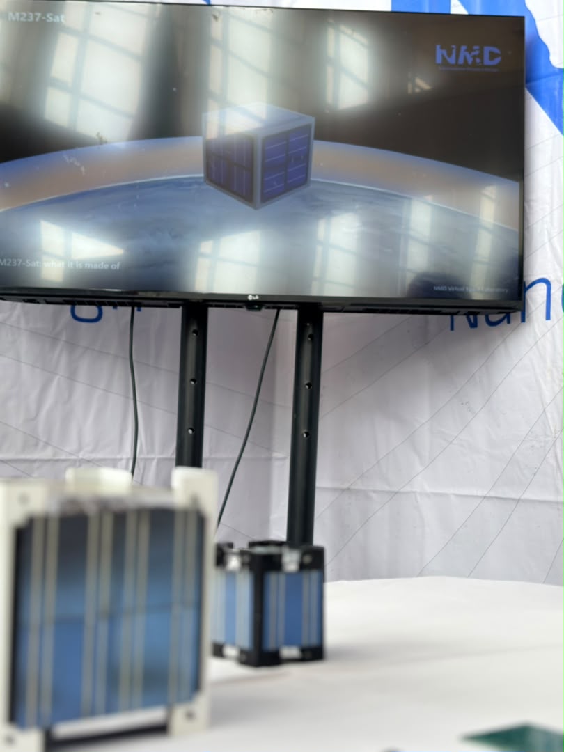 A screen showing the M237-Sat design in the NMD Virtual Space Laboratory, with the real satellite models on the table in front of it